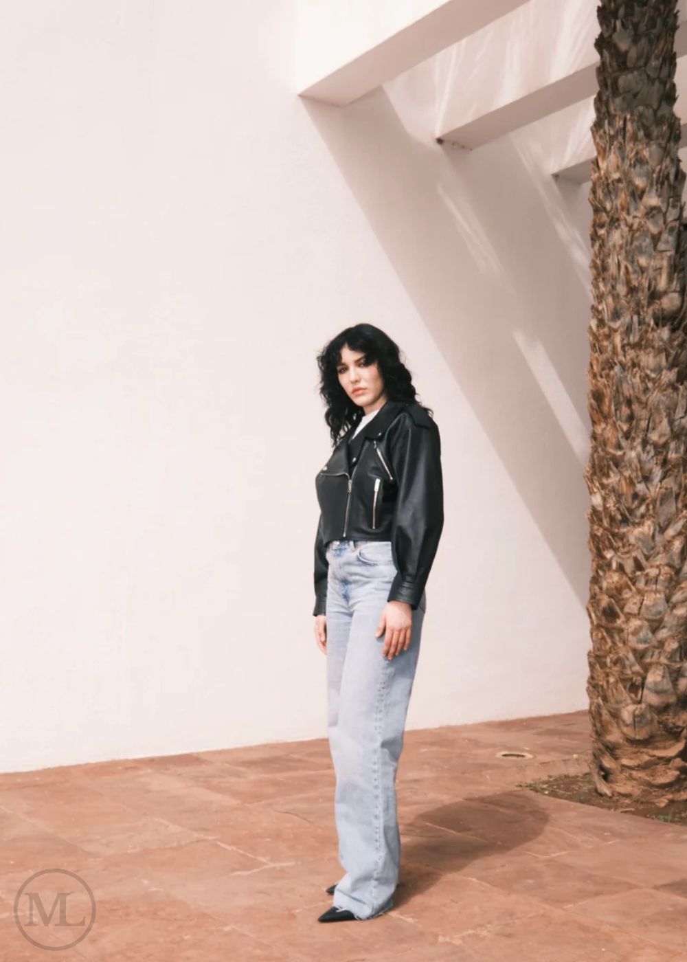Women wearing a Cropped black leather Biker jacket and light blue jeans standing against a neutral wall with palm trees.